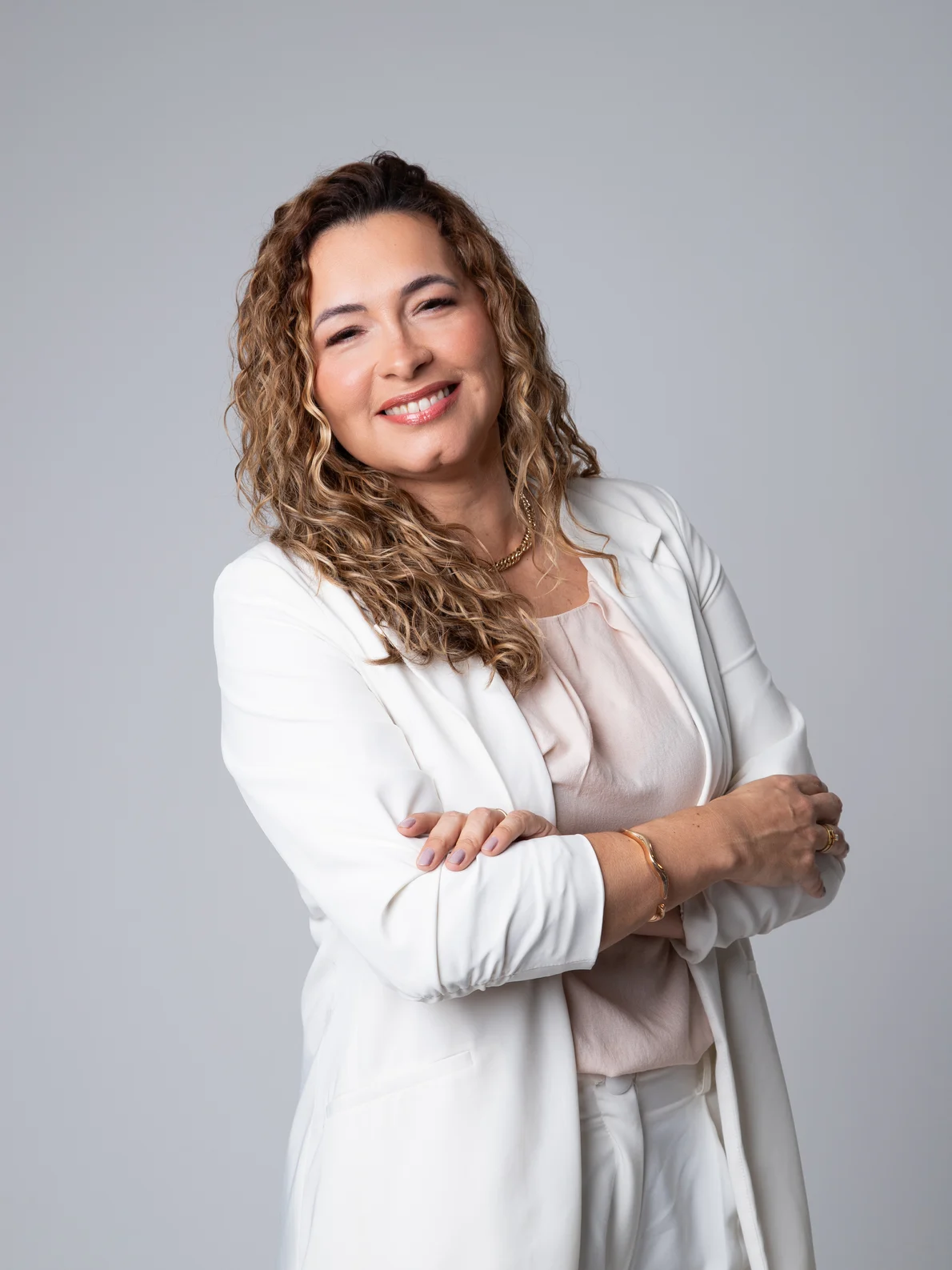 Ádama Nicacio sorrindo, de braços cruzados, blazer branco e cabelo cacheado